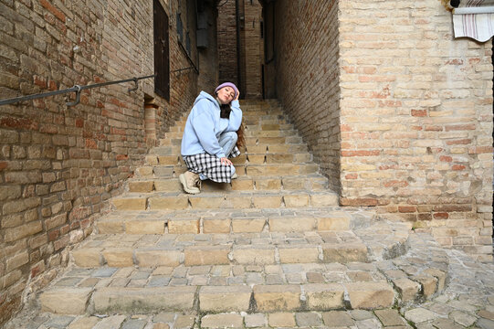 A young woman wearing a blue hoodie and checkered pants sits thoughtfully on old stone stairs of a historic Italian town, surrounded by warm brick walls.