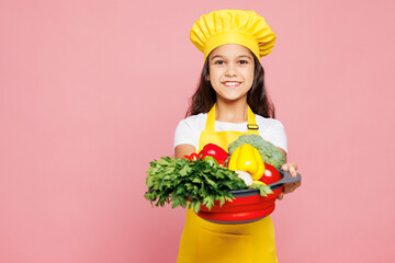 Small kid child girl housewife chef cook baker mother's helper wear yellow apron toque cap hold give bowl with raw vegetables ingredients isolated on pastel plain pink background Cooking food concept