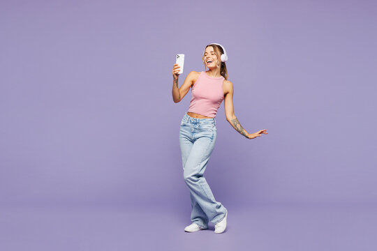 Full body young happy woman wear pink tank top casual clothes listen to music in headphones use mobile cell phone isolated on plain pastel light purple background studio portrait. Lifestyle concept.