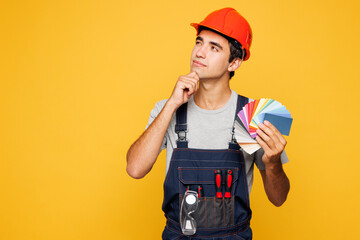 Young minded employee laborer handyman man wear helmet overall hold color palette swatches isolated on plain yellow background. Instruments accessories renovation apartment room. Repair home concept.