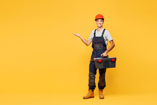 Full body young young employee laborer handyman man he wear helmet overall hold toolbox point aside isolated on plain yellow background. Instrument for renovation apartment room. Repair home concept.