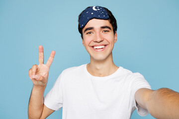 Close up calm young man wear pyjamas jam sleep eye mask rest relax at home doing selfie shot pov on mobile cell phone show v-sign isolated on plain pastel blue background. Good mood night nap concept.