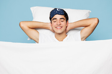 Calm young man he wear pyjamas jam sleep eye mask rest relax at home lies wrap covered under blanket duvet close eyes isolated on plain pastel light blue cyan background. Good mood night nap concept.