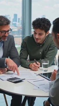 Vertical shot of a financial advisor or manager pointing to a document to explain terms to a young client during a business meeting, with a calculator on the table and a city view.