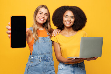 Two young friends buddies IT women wear denim casual clothes together hold work on laptop pc computer use blank screen mobile cell phone isolated on plain yellow orange background. Lifestyle concept.