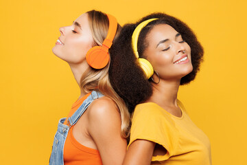 Close up two young friends buddies women wear denim casual clothes together listen to music in headphones dance have fun isolated on plain yellow orange background studio portrait. Lifestyle concept.