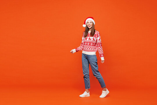 Full body side view merry smiling young woman wear red knitted Christmas sweater Santa hat posing walk go look camera isolated on plain orange background. Happy New Year celebration holiday concept.