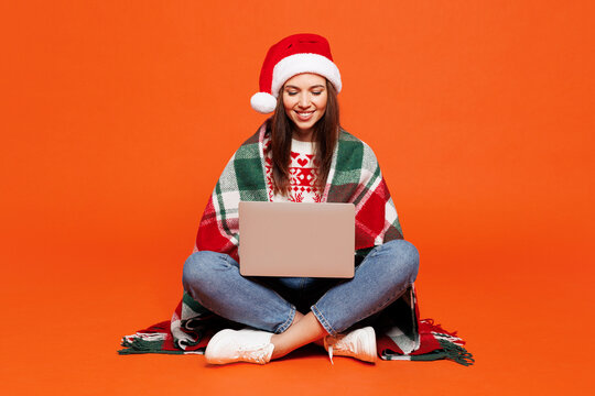 Full body merry young IT woman she wear warm plaid red Christmas Santa hat posing hold use work on laptop pc computer isolated on plain orange background. Happy New Year celebration holiday concept.