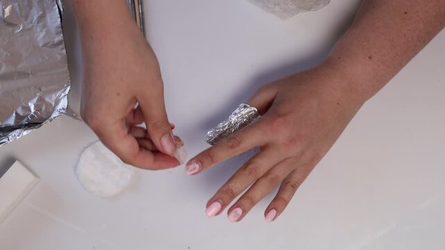 Close-up of hands performing a gel nail removal process by placing soaked cotton pads on nails and wrapping them in foil on a clean white surface
