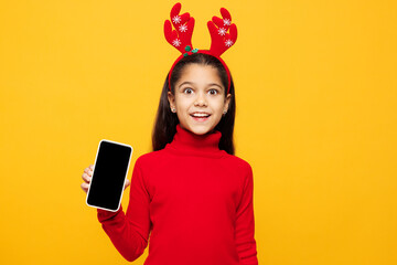 Naklejka premium Merry little girl wear red clothes fun decorative deer horns on head posing hold use blank screen mobile cell phone isolated on plain yellow background. Happy New Year Christmas celebration concept.