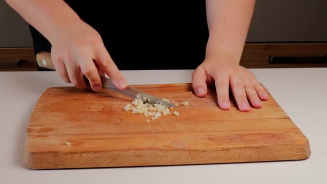 Close-up of hands mincing garlic cloves into small pieces with a knife on a wooden board during home cooking preparation