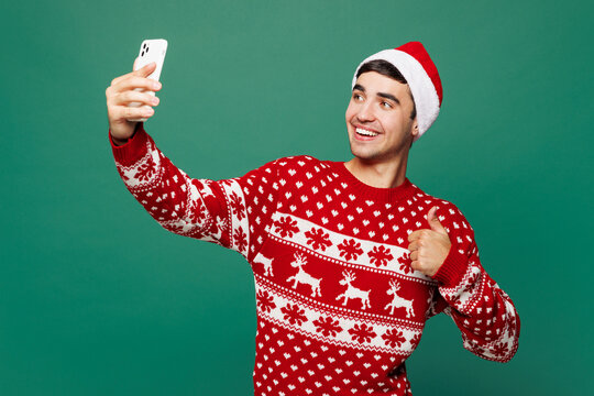 Merry young man wear red warm cozy Christmas sweater Santa Claus hat posing doing selfie shot on mobile cell phone show thumb up isolated on plain green background. Happy New Year celebration concept.