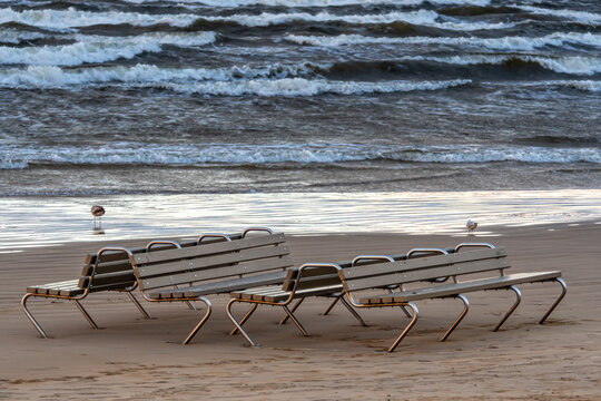 An empty bench sits on sandy beach as ocean waves roll in under a dramatic cloudy sky.