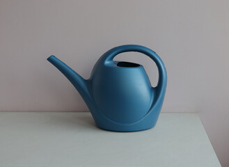 Blue watering can on a white surface with a pink wall in the background. A blue watering can sits on a white surface, perfect for gardening and watering plants indoors or outdoors