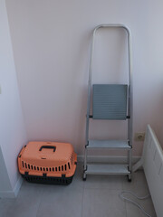 Orange pet carrier and step ladder against a pink wall in a room. A pet carrier and a step ladder are positioned in a corner of a room, next to a wall