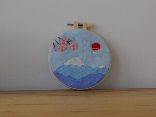 Handmade felt art of Mount Fuji with cherry blossoms and a red sun. This charming piece of fiber art depicts a serene Japanese landscape with a mountain and blooming flowers