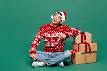 Full body merry young man wear Christmas sweater Santa Claus hat posing sit hold point finger on present box with gift ribbon bow look aside isolated on plain green background. Happy New Year concept.