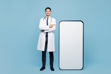 Full body young male doctor man in white medical gown suit work in hospital clinic office stand near big huge blank screen area mobile cell phone smartphone isolated on plain blue background studio