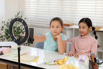 girls are using a smartphone and a ring light to film an origami tutorial video. Colored paper and finished paper crafts are on the desk, showcasing the creation of online 