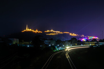 nighttime photograph of Khao Wang (Phra Nakhon Khiri) in Phetchaburi, brightly illuminated by festival lights. Railway tracks run across the foreground with light trails from a moving train,