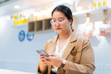 Young woman with glasses using smartphone in modern indoor space, wearing beige blazer and white shirt, focused expression, casual technology interaction