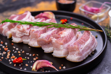 Fresh pork belly arranged on a black plate with spices and herbs in a kitchen setting during the day