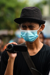 Asian young man filming with Video Camera Outdoors Wearing Protective Mask. Concept of journalism, documentation, health awareness, and public reporting during pandemic.