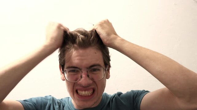 A stressed, angry, and frantic young man pulls his own hair, screams, and grinds his teeth in frustration and despair.