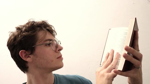 A young male student or nerd with glasses reads a book intently, turning the page. Education and learning concept.