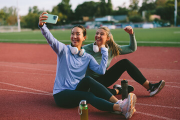 Diverse female friends and athletes, one Asian and one Caucasian, take a joyful selfie on a running track with one friend flexing her fist in a sign of fitness triumph or success