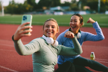 Two diverse female friends and athletes proudly pose, flexing their biceps for a triumphant selfie photo after a successful and fun workout on a running track