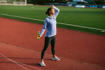 Exhausted Asian female athlete wipes sweat from her brow with relief, taking a hydration break on a running track after a strenuous and intense outdoor workout