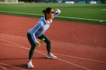 Focused Asian female athlete takes a quick break on a running track, shielding her eyes from the sun or wiping sweat while holding a sport drink bottle after an intense run