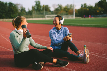 Two diverse female friends and athletes, one Asian and one Caucasian, share a joyful laugh and look at a mobile phone during a relaxed hydration break on a running track