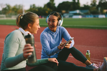 Excited Asian female athlete shares shocking or funny content on her mobile phone with her Caucasian friend during a hydration break on an outdoor running track