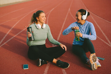 Diverse female athletes, one Asian and one Caucasian, take a joyful hydration and conversation break, sitting on a running track and drinking from their sport bottles after a workout