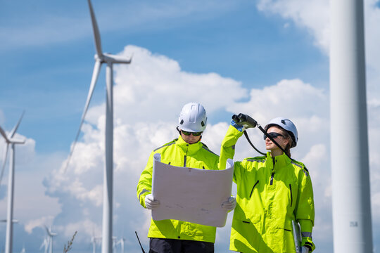 Two men in yellow jackets are looking at a piece of paper. One of them is holding a pair of binoculars. Engineers on wind turbine farm, inspecting and planning for renewable energy project. - Powered by Adobe