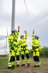 Three people in yellow and black safety gear are standing in front of a wind turbine. Group of engineers with PPE, collaborating and walking at wind turbine field, concept of achievement.