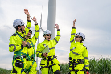 A team of workers in bright yellow safety gear are gathered around a large wind turbine. Group of engineers with PPE, collaborating and walking at wind turbine field, concept of achievement.