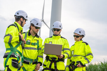 A group of four workers in yellow safety gear are gathered around a laptop. Group of engineers (four people) with PPE, collaborating and walking at wind turbine field, concept of achievement.
