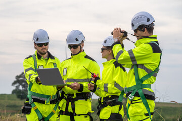 Four men in yellow and green safety gear are looking at a laptop. One of the men is holding a camera. Technicians in PPE discussing blueprint and inspecting wind turbines, planning for maintenance.