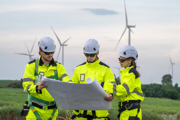 Three workers in bright yellow and white safety gear are examining a blueprint. Technicians in PPE discussing blueprint and inspecting wind turbines, planning for maintenance.