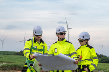 Three people in yellow and white safety gear are looking at a drawing of a wind farm. Technicians in PPE discussing blueprint and inspecting wind turbines, planning for maintenance.