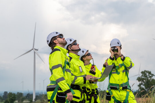 Four people wearing yellow and green safety gear are standing on a hill looking at a wind turbine. Technicians in PPE discussing blueprint and inspecting wind turbines, planning for maintenance.