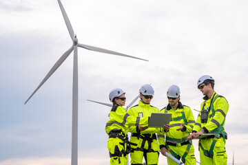 Four people are standing on a wind farm, looking at a laptop. They are wearing safety gear. Technicians in PPE discussing blueprint and inspecting wind turbines, planning for maintenance.