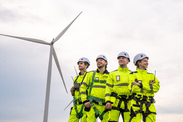 Four workers in bright yellow and green safety gear are standing in front of a large wind turbine. Technicians in PPE discussing blueprint and inspecting wind turbines, planning for maintenance.