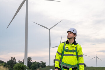 A man in a yellow jacket stands in front of a wind farm. He is wearing a hard hat and a harness. Wind turbine specialist inspecting site plans, ready for working at height with safety equipment.
