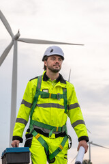 A man in a yellow and green safety suit is holding a blue toolbox. He is wearing a hard hat. Wind turbine specialist inspecting site plans, ready for working at height with safety equipment.