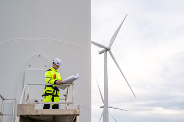 A man is standing on a platform next to a wind turbine. He is wearing a yellow jacket and a hard hat. Wind turbine specialist inspecting site plans, ready for working at height with safety equipment.