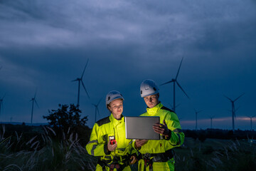 Two workers in yellow jackets and hard hats are examining a laptop. Technician team in safety harnesses, inspecting wind turbines with technology at twilight.